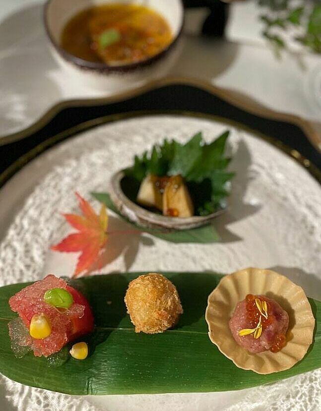 Đĩa khai vị tổng hợp gồm bánh tart cá ngừ vây xanh, khoai lang cá tuyết chiên giòn, trái cây thạch tảo bẹ, bào ngư hấp xốt gan. Các món ăn được bày trên một chiếc lá cây thuộc họ tre trúc, bên cạnh là một chiếc lá phong đỏ, mang phong cách Nhật Bản.